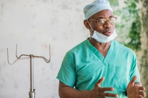 Real people. Male doctor wearing scrubs, glasses, and face mask standing and speaking in medical facility in Liberia.