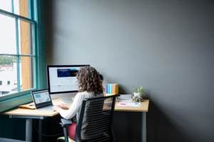 Female office worker sitting at desk using Surface Laptop in casual office setting.