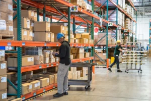 Male worker wearing hardhat using tablet in warehouse. Another worker in background.