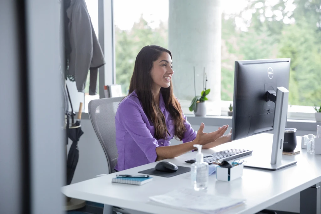 A female employee talking on a video call with hand sanitizer on her desk, socially distancing from others. Hybrid Workplace collection.