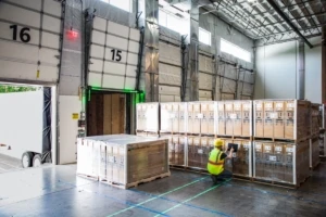 Male worker in hardhat and reflective vest using tablet in warehouse loading dock. Tablet screen not visible.