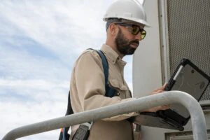 Field engineer viewing data on a laptop after inspecting turbines on a wind farm. Keywords: Dynamics 365; operations; green energy; outside; outdoors; on the go; remote assist; training; man; wearing hard hat and safety gear