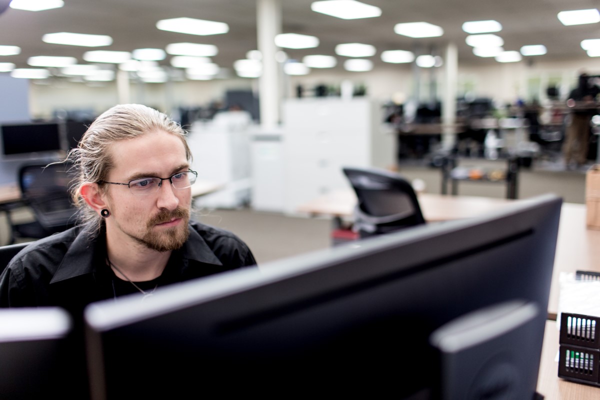 Male worker sitting at desk in manufacturing office and looking at two monitors (screens not shown).