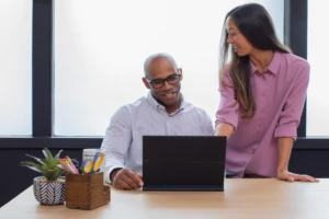 Man sitting at desk in a small business office with an HP Elite Folio, which is a commercial device, collaborating with woman standing.