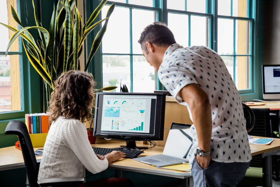 A woman sitting at a desk using a computer next to a man standing, looking at the screen.