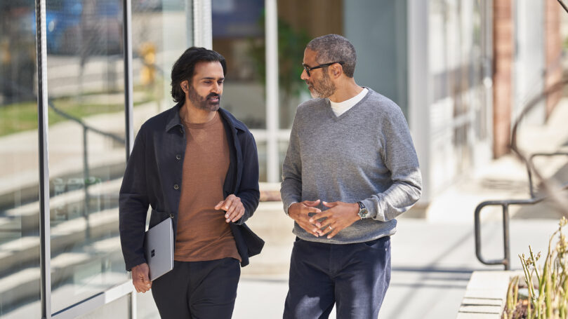 Two people walking and talking, one of them is holding a laptop.