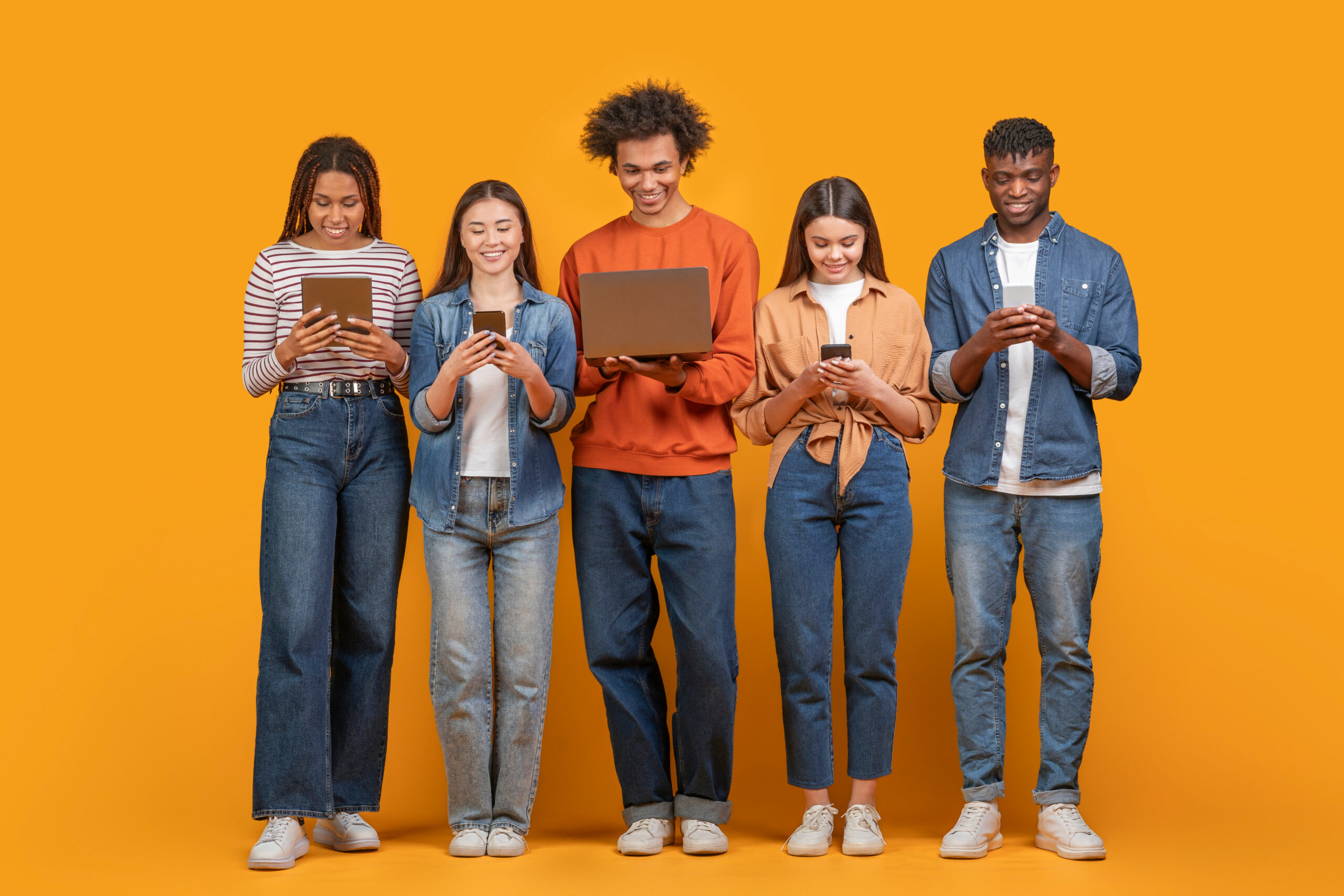 This image shows a group of five young adults standing in a row against a yellow background. They are all looking down at their devices, which include a tablet, a smartphone, and a laptop