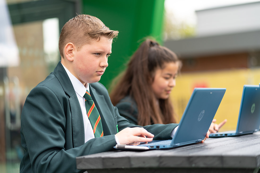 Two high school students sitting at a table in school, working on their laptops.