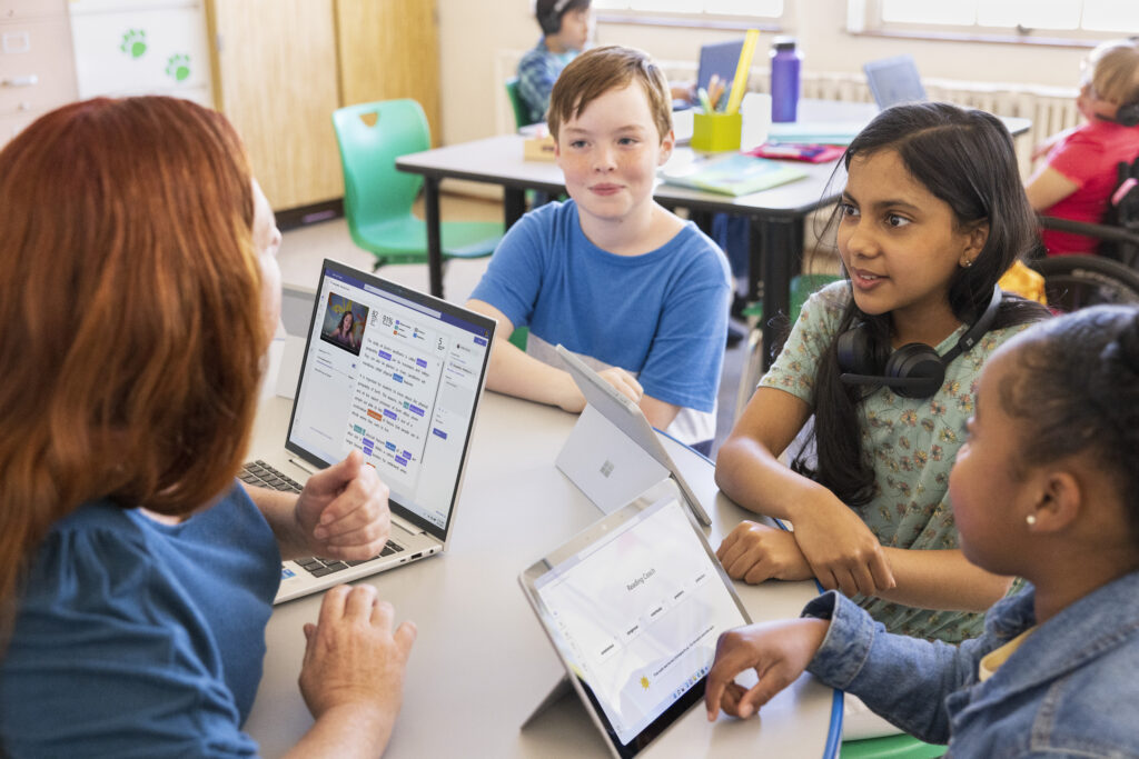 A teacher working with three elementary school students in a classroom setting, using AI tools on a laptop and tablets. 