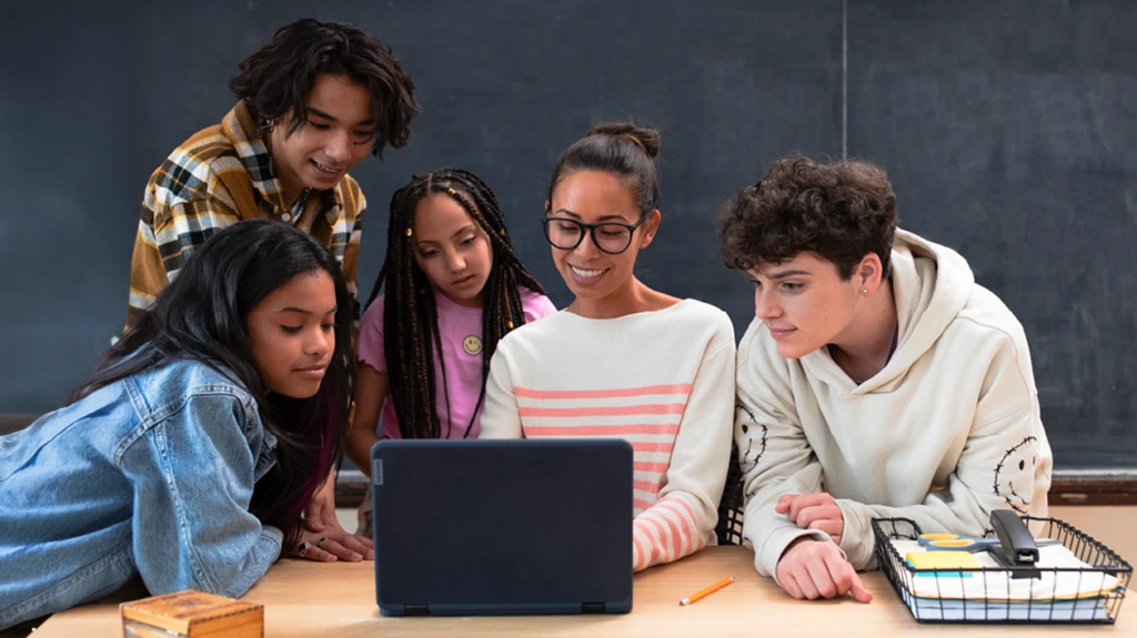 An educator and four students looking at a laptop at the educator’s desk in a school classroom.