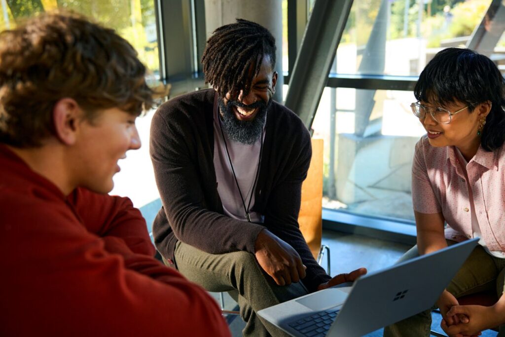 An instructor and a group of students collaborate on a laptop in a common area at a college.