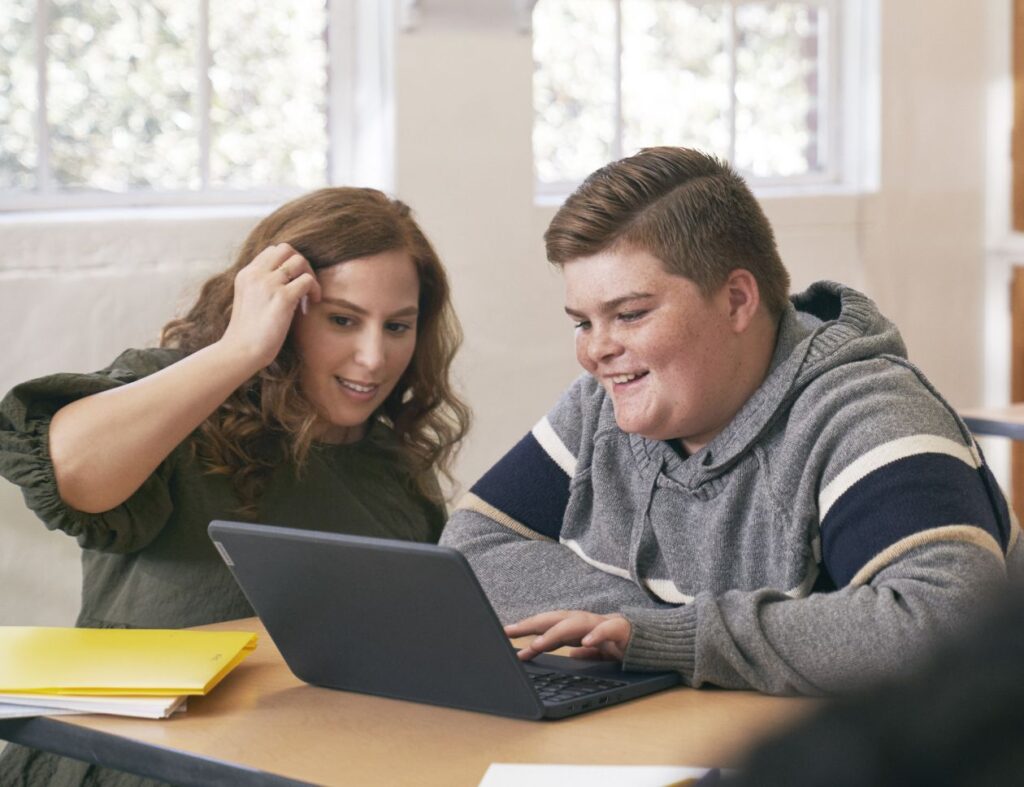 An educator kneeling at a student’s desk and helping him with schoolwork on his laptop.