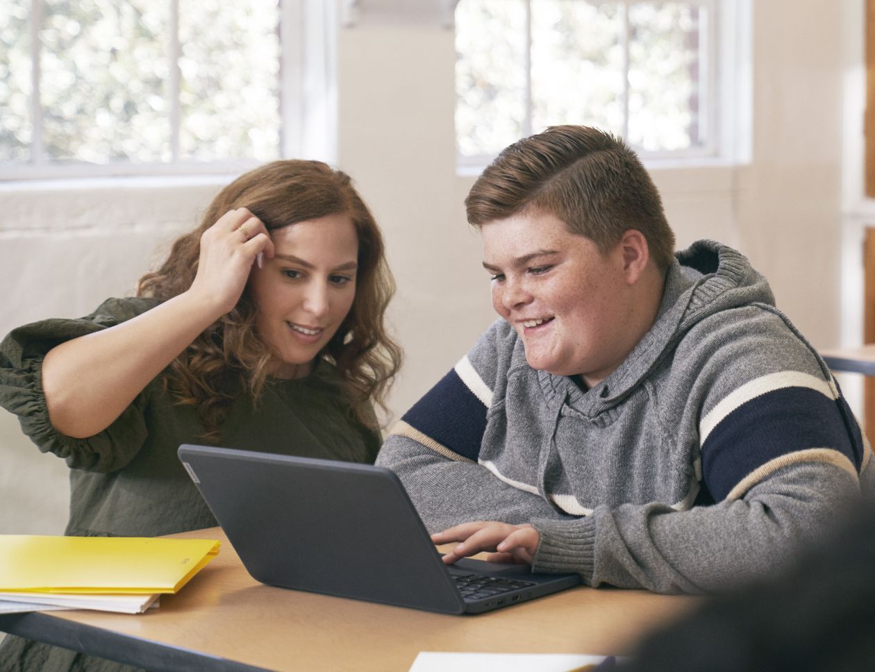 An educator kneeling at a student’s desk and helping him with schoolwork on his laptop.