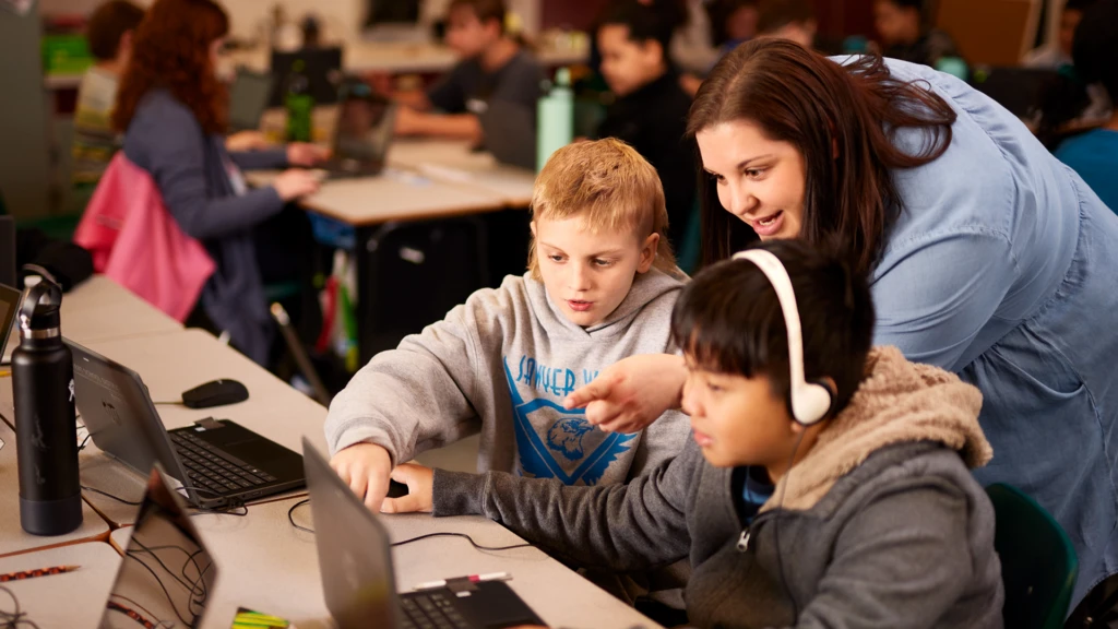 A teacher speaking to two elementary school students in a classroom setting, who are using Minecraft Education on laptops.