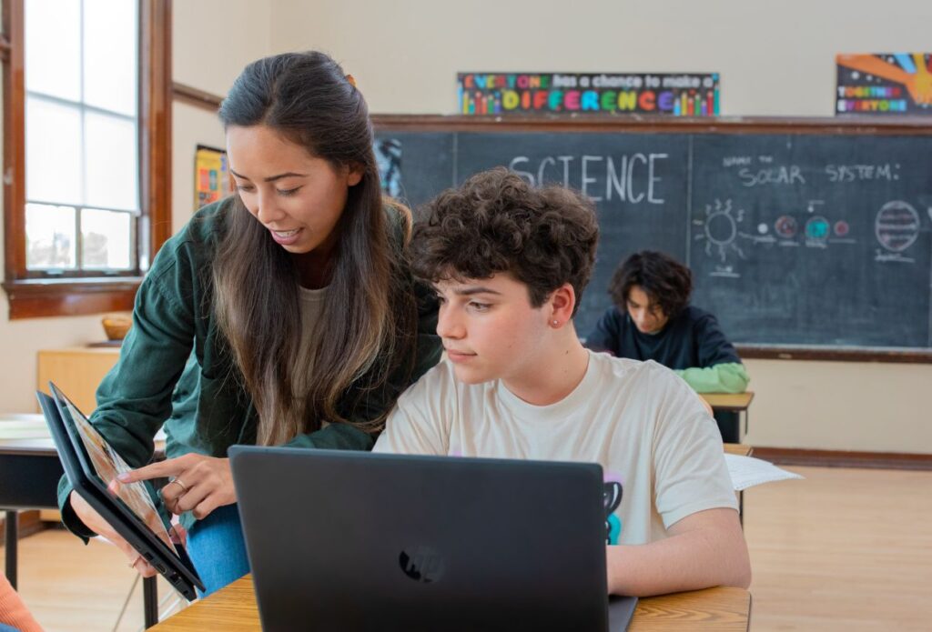 A teacher engages with a student at his desk. They're both using laptops.