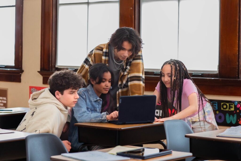 Four students gathered around an open laptop at a desk in a school classroom.