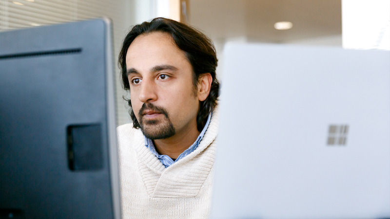 Male office worker looking at monitor on desk (screen not shown). A open Surface Laptop is also on his desk (screen not shown)