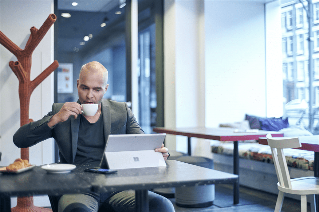 Picture of a man drinking coffee at a cafe looking down at a Microsoft tablet.