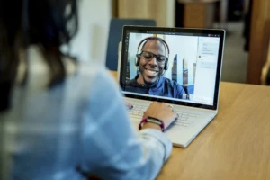 Woman at a desk using a Surface laptop to make a Microsoft Teams video call with one man smiling and wearing a headset. Business Voice conference call/meeting device is in the background.