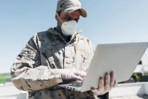 a close up of a soldier in camo wearing a mask and holding a laptop