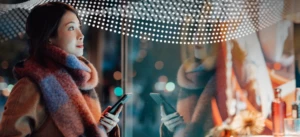 Woman holds cell phone in front of retail storefront display with merchandise at night.