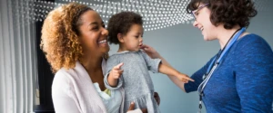 Smiling woman with brown skin holds a small child while engaged in conversation with a female clinician and the small child points at the female clinician.