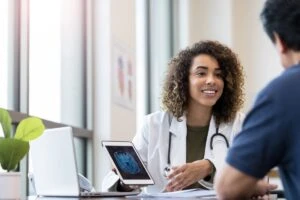 Female clinician w/ brown skin and brown shoulder length curly hair shows computer screen with unclear image of results to patient.