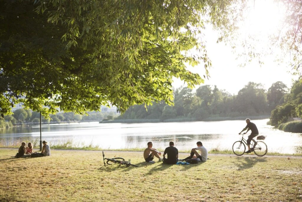 Two groups of 3 women and 3 men sitting in a park next to a lake. Also a man riding his bike along a path around the lake