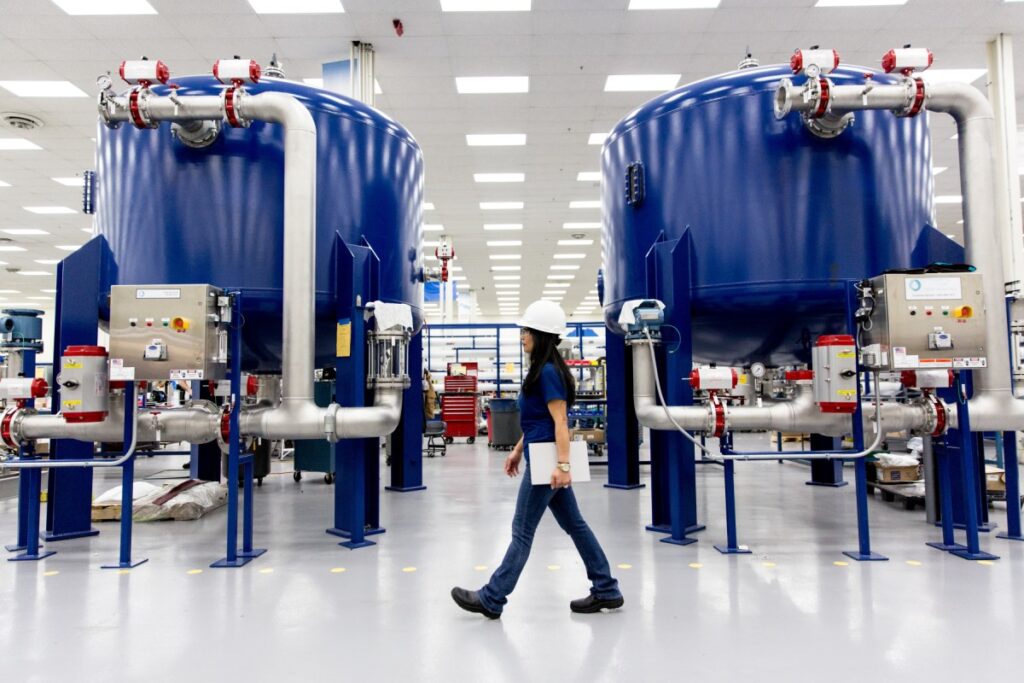 Female worker walking along factory floor in manufacturing plant, in front of two large blue vats. She is wearing a hardhat and safety glasses, and holding a closed tablet by her side.