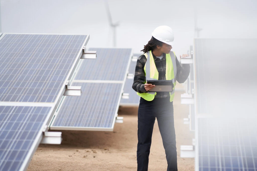 Field engineer inspects solar panels on a wind farm using remote assist on a Surface tablet while wearing a reflective safety vest and a hardhat.