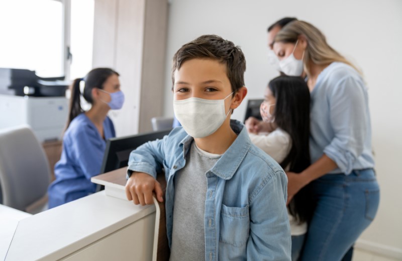 Photo of a young patient with a mask on at looking happy in the foreground. The rest of the family in the background check-in with the office manager.