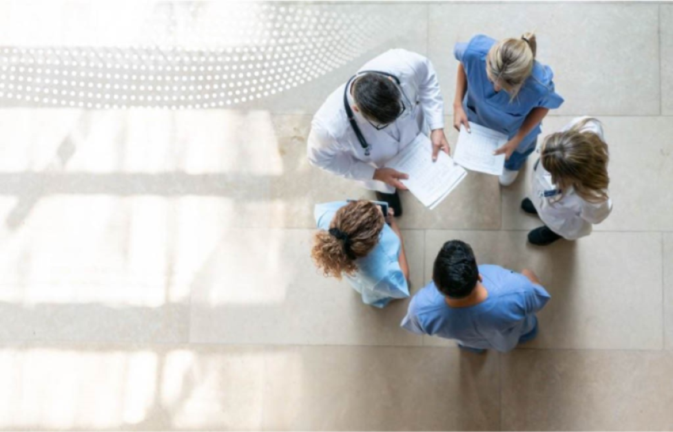 An overhead shot of a group of medical professionals standing in a circle in conversation on a beige tiled floor.