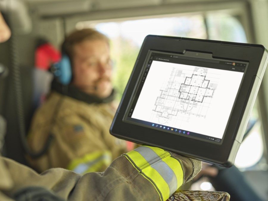 In the forefront, the arm of a firefighter sitting in the passenger seat of a firetruck holding a Surface tablet to look at blueprints of a building and a male firefighter in the background in the driverâs seat with a headset on.
