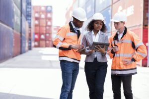 2 males and 1 female standing at a port with hard hats on looking at an tablet device