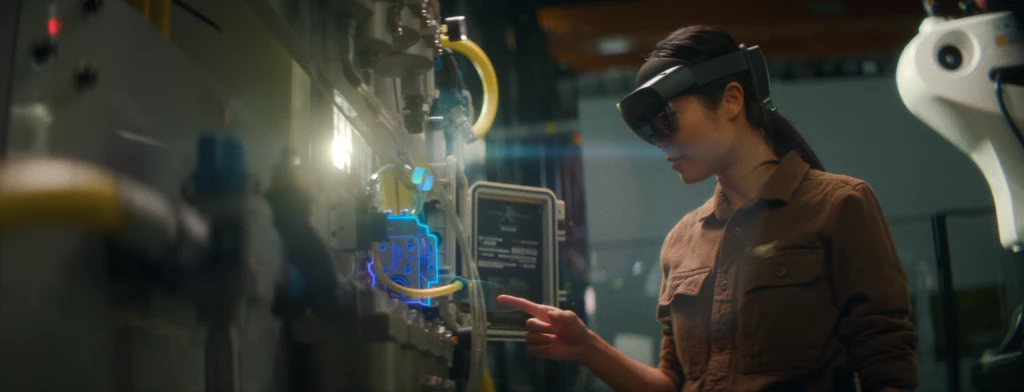 Woman in a HoloLens 2 gestures with her index finger at a machine. The part she points to is lit up with a holographic outline.  