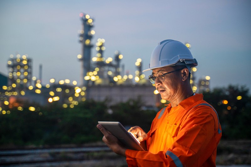 An engineer wearing a hardhat uses a digital tablet while working the night shift at petroleum oil refinery.