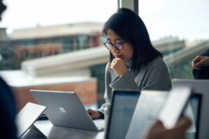 Woman with glasses sitting at a community table in common area reading on a Surface laptop