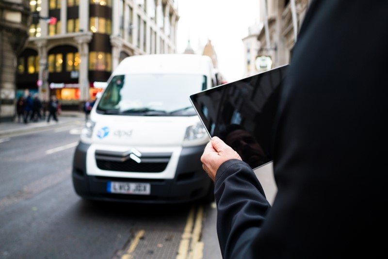 Male commuter holding tablet on city street sidewalk. His reflection is partially visible on blank tablet screen. A white van is driving on street in foreground; pedestrians walking on sidewalk in background.