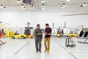 Two male first line workers in conversation while walking among assembled helicopters on factory floor in commercial manufacturing plant.
