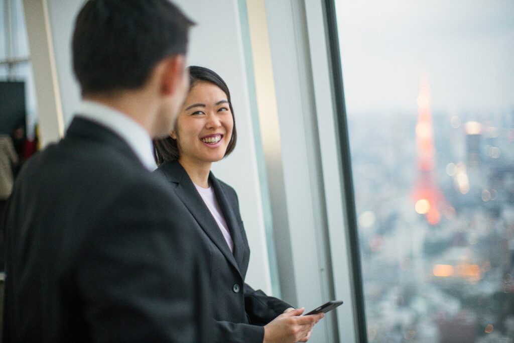 A woman and man, both wearing business attire, stand in front of a window in a tall building. She holds a phone and is smiling.