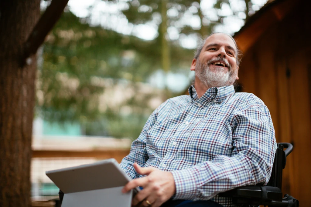 Stuart Pixley, a man who uses a wheelchair, smiles while holding a laptop on his lap outside.