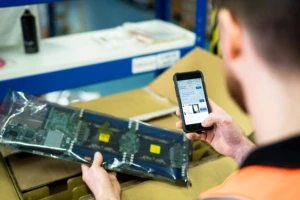 Man wearing an orange vest in a manufacturing or warehouse facility who is taking a picture of a product and messaging about it on his phone in Microsoft Teams chat. As we return to work, we will increasingly use technology to connection and collaborate.