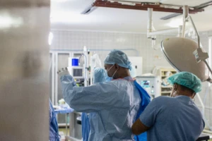 A medical worker helps a male surgeon into medical scrubs in operating room in medical facility.