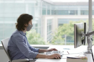 A man in a facemask sitting in front of a window on a computer. Analytics can be used for customer insight.