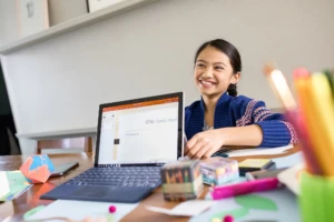 A child doing remote learning. She is sitting by a table with a computer and stationary around her.