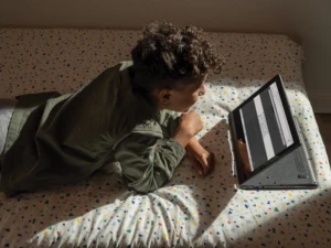 A child doing remote learning. He is laying on a bed reading his computer.