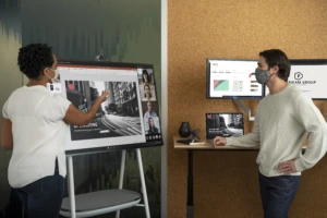 Two adults in face masks collaborating on a PowerPoint presentation in an office while using a Microsoft Surface Hub 2S 50” device during a Teams video call.