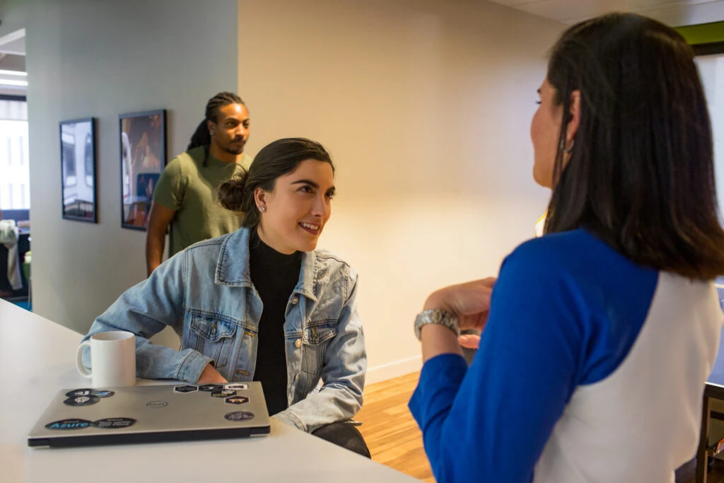 Real people, real offices. Female developer collaborating with a colleague in office breakroom or kitchen. Colleagues play ping pong in the background. One developer has personalized her PC laptop with stickers.