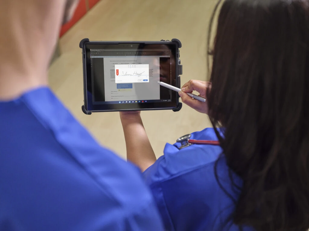A nurse holds a Surface Go 3 in a ruggedized case in one hand a Surface Pen in the other getting ready to sign a document with AdobeSign on the screen.
