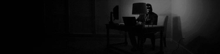 A black and white photo of a person sitting behind a computer, wearing a mask